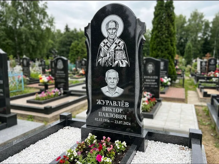 Памятник с гравировкой иконы: стела с образом в полный рост на обороте — гравировка образа Матроны Московской, низкие кусты спиреи в цвету, работа «Лебедева» в Волгограде