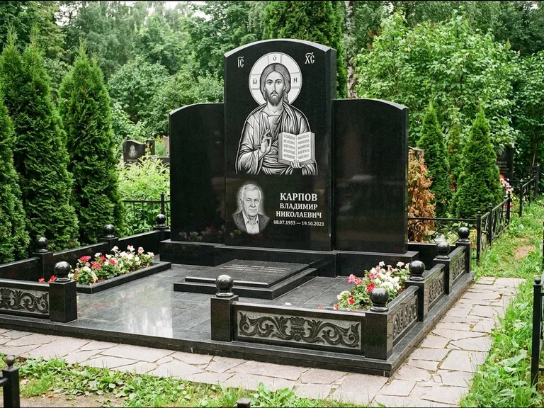 Памятник с гравировкой иконы: широкая панорамная стела с золочёным нимбом — гравировка образа Матроны Московской, цветник с белыми хризантемами, работа «Мельникова» в Волгограде
